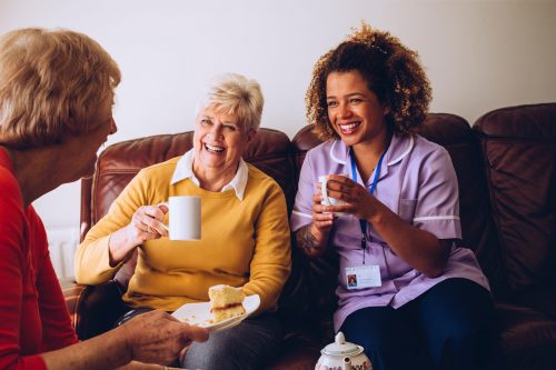 Elderly carer sitting with two of her patients in the care home. They are enjoying some cake and tea.